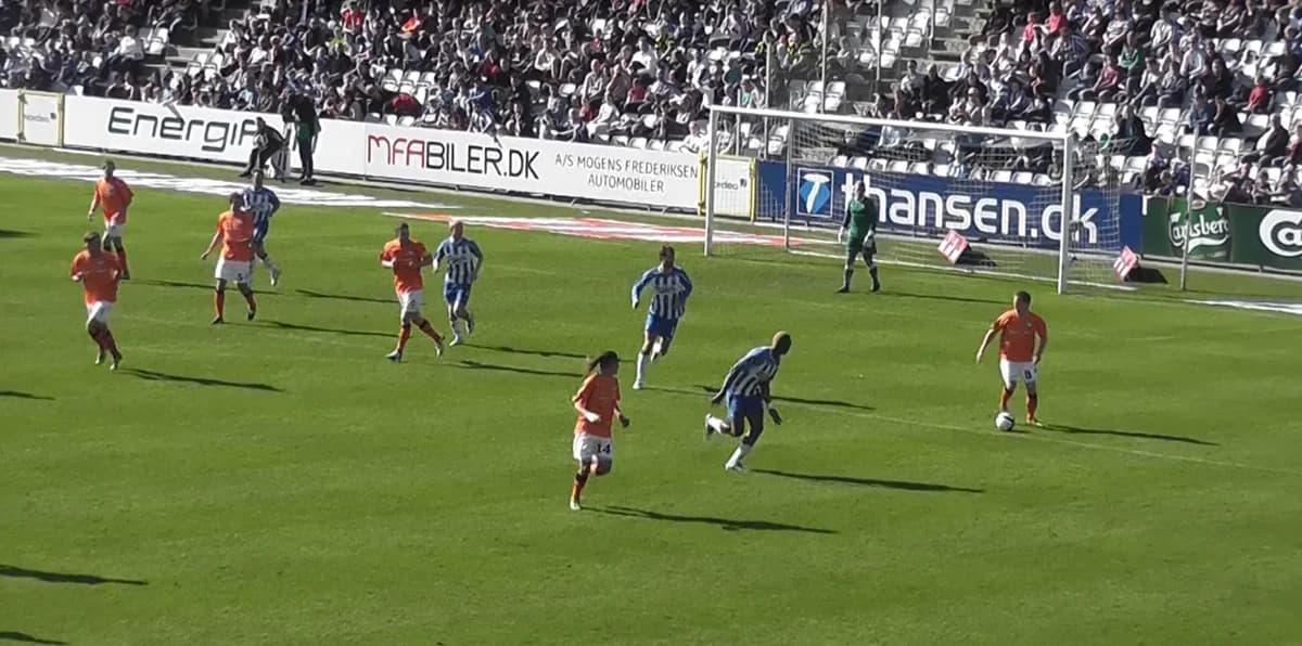 Football match between teams in orange and blue/white stripes with a goalkeeper in green