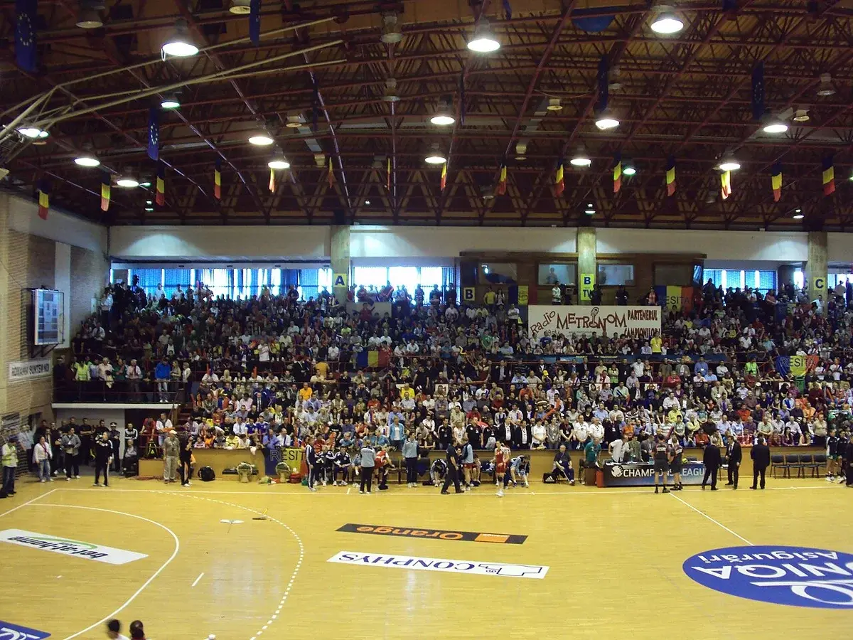 A crowded indoor sports arena with a handball court, hanging flags, and banners above the stands.