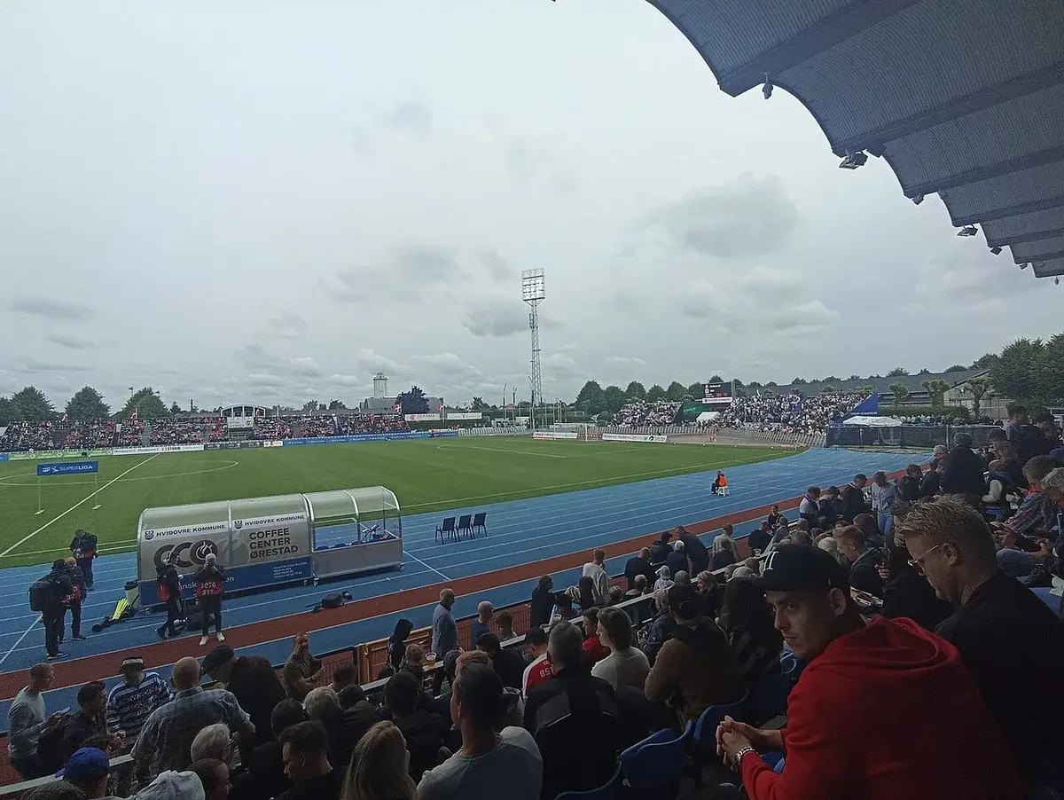 Wide view of a football stadium with spectators, a blue running track, and an away supporters’ section.