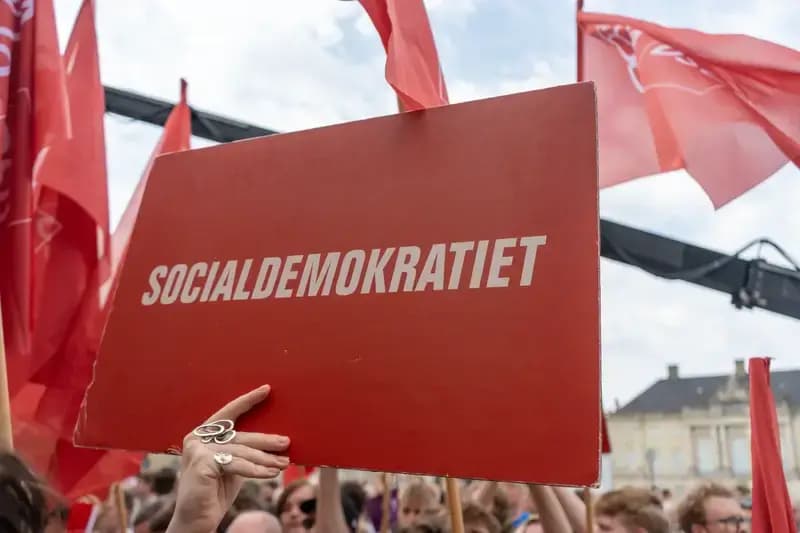 Person holding Socialdemokratiet sign at outdoor event with red flags