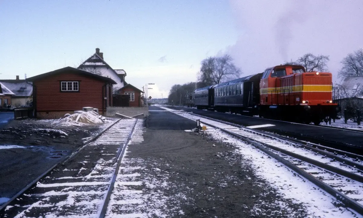 A red diesel locomotive pulling two passenger coaches past a snowy station platform.