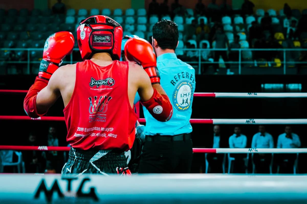 A Muay Thai boxer in red gear stands in the ring next to a referee.